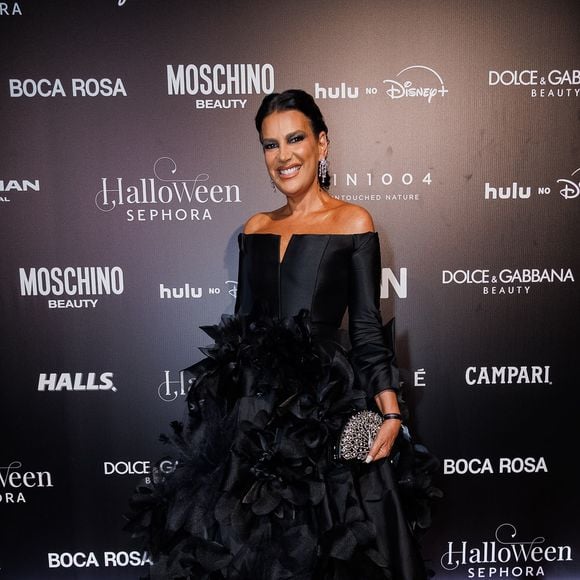 Andréia Guimarães veste um vestido de gala preto dramático com decote ombro a ombro estruturado e fenda central discreta no busto. A peça se destaca pela saia volumosa e rodada rica em aplicações tridimensionais de grandes flores e texturas em relevo por toda a extensão, combinada com clutch prateada rendada, brincos pendentes e cabelos presos num coque polido