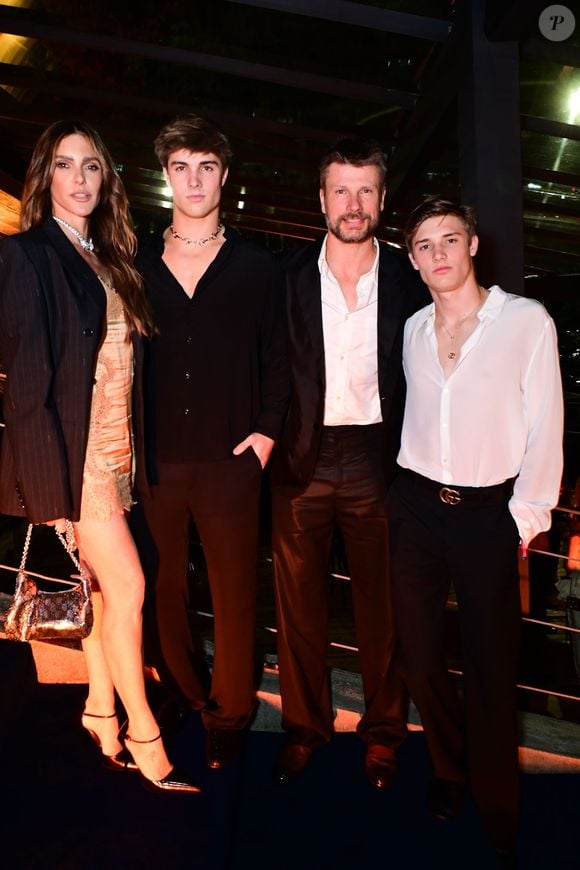 Fernanda Lima veste um minivestido acetinado em tom nude/dourado com detalhes em renda, sobreposto por um blazer oversized preto com risca-de-giz. Completa com meia-calça, sapatos de salto e bolsa prateada com alça de corrente; Rodrigo Hilbert usa um terno preto clássico de corte reto combinado com camisa social branca de gola aberta e sem gravata; João e Francisco: um veste camisa preta fluida de gola V com calça preta e colar de corrente; o outro usa camisa social branca levemente translúcida por dentro de calça preta com cinto de fivela prateada