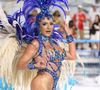 O figurino é dominado por tons de azul safira e azul royal, aplicados em pedrarias de alto brilho que cobrem busto, quadris e acessórios. O destaque central é o cocar vertical exuberante, estruturado com plumas longas em camadas, que se abrem em leque acima da cabeça e criam uma silhueta alongada