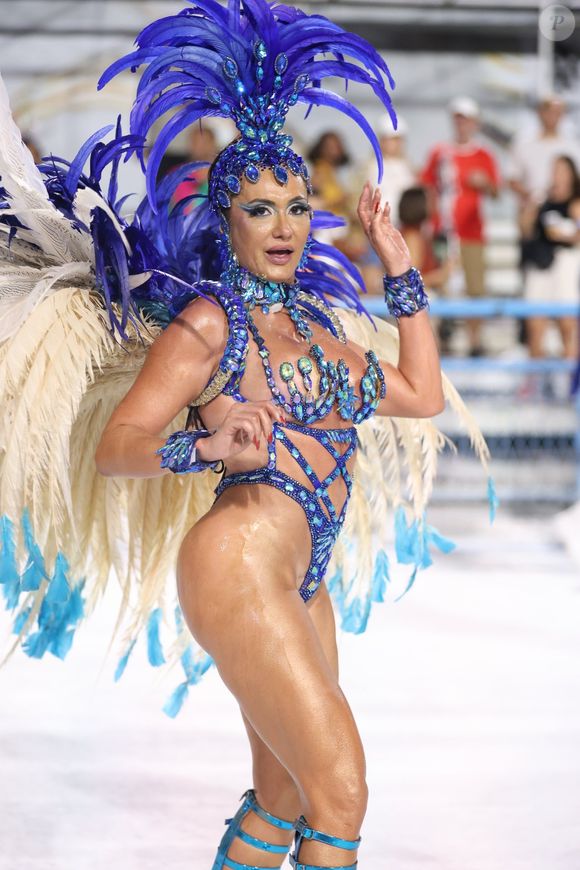 O figurino é dominado por tons de azul safira e azul royal, aplicados em pedrarias de alto brilho que cobrem busto, quadris e acessórios. O destaque central é o cocar vertical exuberante, estruturado com plumas longas em camadas, que se abrem em leque acima da cabeça e criam uma silhueta alongada