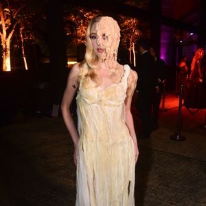 Carrie, a Estranha + A Substância! Fefe Schneider veste um vestido longo branco de alças finas com saia esvoaçante repleta de fendas e recortes que criam um efeito de tiras esfarrapadas. O grande destaque é o lado direito do rosto da modelo, coberto por uma prótese que imita cera derretida ou teias de aranha em tons de bege, estendendo-se do cabelo até o queixo e cobrindo um dos olhos. Ela também possui uma pequena tatuagem de borboleta no braço direit
