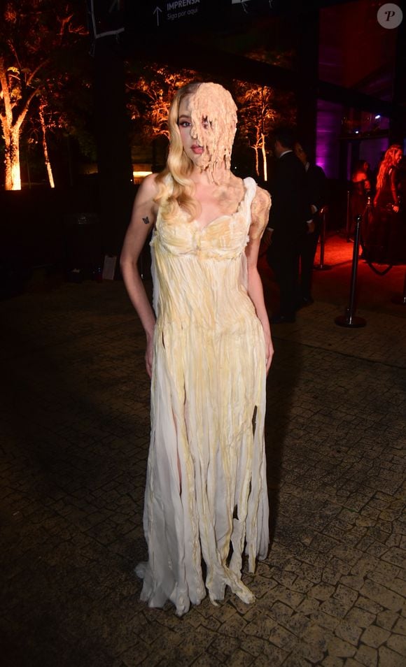 Carrie, a Estranha + A Substância! Fefe Schneider veste um vestido longo branco de alças finas com saia esvoaçante repleta de fendas e recortes que criam um efeito de tiras esfarrapadas. O grande destaque é o lado direito do rosto da modelo, coberto por uma prótese que imita cera derretida ou teias de aranha em tons de bege, estendendo-se do cabelo até o queixo e cobrindo um dos olhos. Ela também possui uma pequena tatuagem de borboleta no braço direit