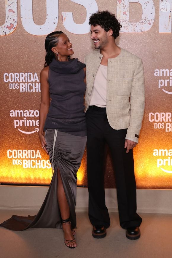 Em pose conjunta, Thainá Duarte e Matheus Abreu harmonizam produções de moda autoral e contemporânea. Enquanto a atriz exibe a silhueta ajustada e assimétrica em tons de cinza com cauda fluida, o ator complementa a composição com a textura encorpada do tweed claro sobre alfaiataria escura