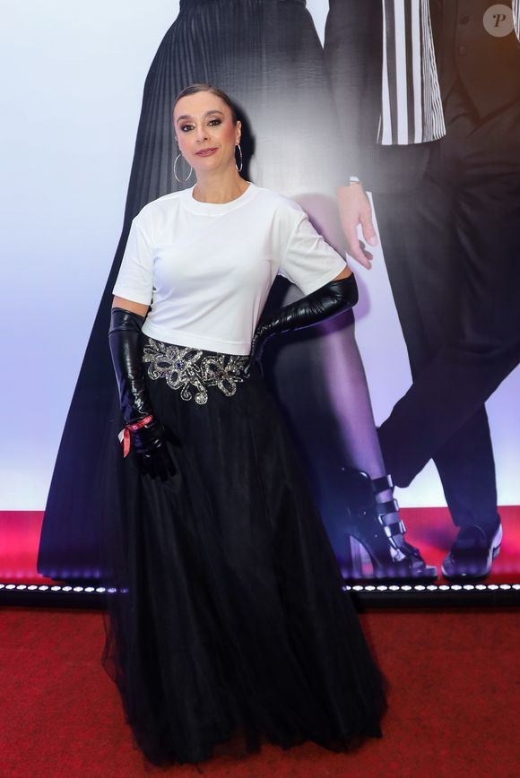 Cátia Fonseca utilizou uma proposta que mescla o básico com o dramático. O look traz uma t-shirt branca de algodão com corte reto, inserida sob uma saia longa de tule preto de várias camadas e cintura alta, decorada com um bordado prateado de motivos orgânicos na região do cós. O elemento de maior impacto são as luvas de ópera em vinil preto que cobrem quase todo o braço, trazendo um ar performático à produção