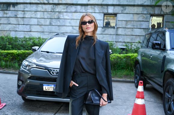 Sasha optou por um conjunto total black composto por um blazer oversized de ombros estruturados sobre uma blusa de gola alta em seda e calças de corte reto impecável, finalizando com óculos escuros estilo cat-eye e uma bolsa de mão estruturada
