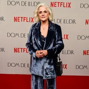 Marieta Severo veste um terno de veludo molhado em tom azul-cinzento de caimento fluido e modelagem ampla. O conjunto é composto por blazer alongado com lapelas clássicas sobre top preto com detalhes em renda no decote, calça reta combinando e botas pretas de cano curto
