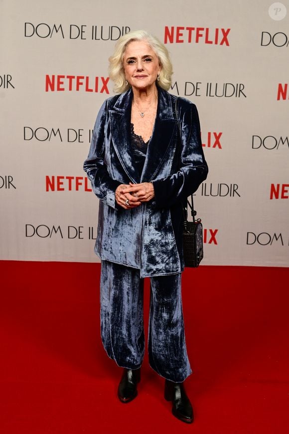 Marieta Severo veste um terno de veludo molhado em tom azul-cinzento de caimento fluido e modelagem ampla. O conjunto é composto por blazer alongado com lapelas clássicas sobre top preto com detalhes em renda no decote, calça reta combinando e botas pretas de cano curto
