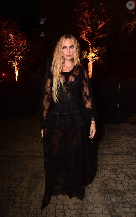Schneider Moura usa vestido longo preto de renda com transparência. A peça apresenta mangas compridas bufantes e transparentes de renda, um corpete ajustado com detalhes de fitas horizontais na cintura e uma saia longa fluida também em renda transparente sobre uma base escura