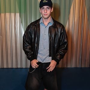 Pietro Antonelli Benício trouxe para o evento a essência do streetwear utilitário com toques de sofisticação. O jovem combinou uma jaqueta de couro preta de modelagem oversized com uma camisa social azul clara levemente desabotoada, criando uma sobreposição texturizada. Na parte inferior, bermudas jeans largas de lavagem escura e meias brancas aparentes com sapatos derby de sola tratorada reforçaram a estética 'indie sleaze'