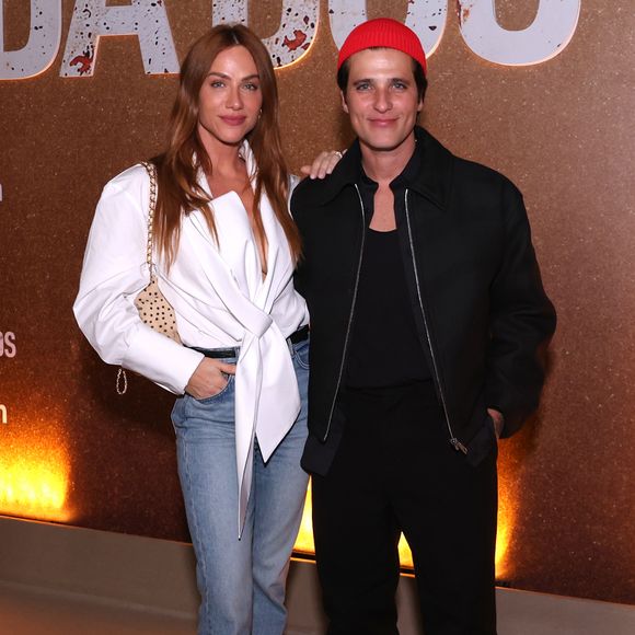 Giovanna Ewbank aposta no contraste hi-lo ao combinar camisa desconstruída branca de modelagem transpassada e decote acentuado com calça jeans reta e scarpin preto de bico fino. Ao seu lado, Bruno Gagliasso mantém a proposta monocromática preta sob jaqueta estruturada de zíper, arrematada pelo gorro vermelho e sapatos Tabi