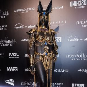 Iza ostenta uma caracterização inspirada no Egito Antigo, especificamente na deidade Anúbis. O corpo está totalmente coberto por pintura corporal preta com brilho. Sobre ela, é usado um conjunto de joias corporais douradas elaboradas e vazadas, incluindo um sutiã estruturado, gola peitoral, braçadeiras, manoplas e cinto com correntes pendentes. O adereço de cabeça inclui as orelhas de chacal e detalhes em ouro e azul. Sapatos de salto alto de bico dourado completam o figurino