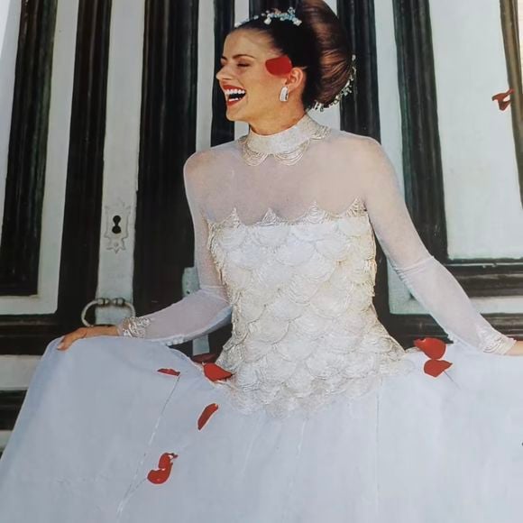 1.Vestido de manga longa em tule

Um clássico das noivas brasileiras do início dos anos 2000. O vestido apresenta uma saia ampla em camadas de tule esvoaçante, com um estilo ousado e romântico. A parte superior tem uma ilusão de transparência com mangas longas em tule, contrastando com bordados em formato de escamas de concha, conferindo brilho e textura. É um look etéreo, quase angelical, mas com um toque dramático pelos detalhes vermelhos.