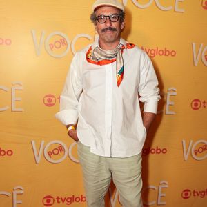 Milhem Cortaz aposta em um estilo descontraído com camisa social branca ampla com mangas dobradas e lencinho estampado amarrado no pescoço, combinado a uma calça de sarja verde-claro com a barra dobrada. A produção é arrematada por boina clara, óculos de grau e tênis esportivos em tom bege