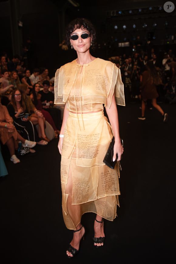 Débora Nascimento personificou o conceito de 'moda-arte' ao surgir com um conjunto de transparências em tom areia. O look é composto por um top cropped e uma saia midi, ambos construídos com sobreposições de retângulos de organza que trazem trechos manuscritos, conferindo uma aura literária e intelectual à produção