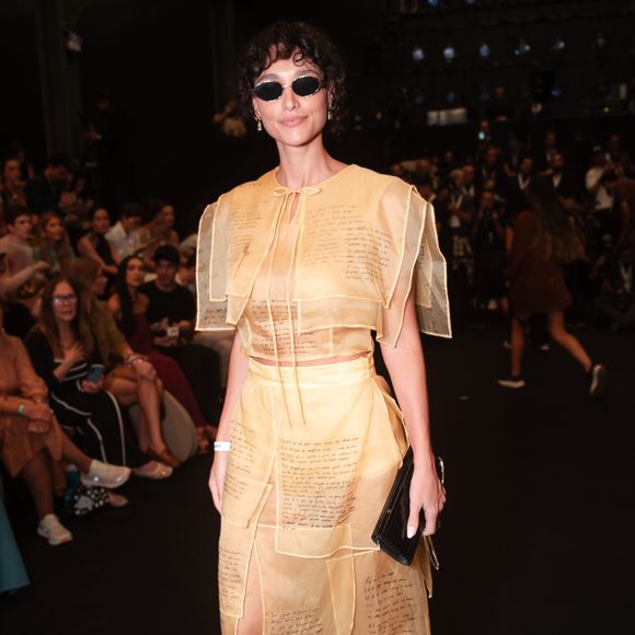 Débora Nascimento personificou o conceito de 'moda-arte' ao surgir com um conjunto de transparências em tom areia. O look é composto por um top cropped e uma saia midi, ambos construídos com sobreposições de retângulos de organza que trazem trechos manuscritos, conferindo uma aura literária e intelectual à produção