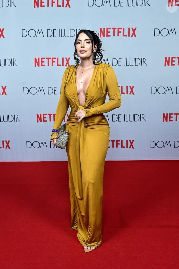 Fernanda Bande veste um vestido longo dourado mostarda em tecido cintilante. O modelo tem mangas longas, decote em V profundo até a cintura e um detalhe de amarração frontal com pontas fluidas que se estendem até a barra. Ela segura uma bolsa de mão Gucci clássica com monograma