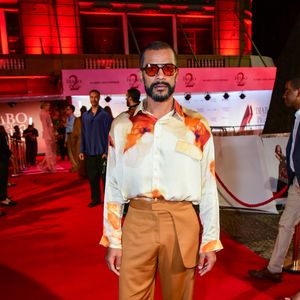 Samuel de Assis trouxe uma proposta de alfaiataria contemporânea e fluida. Ele vestiu uma camisa de seda com estampa abstrata/floral em tons terrosos e fundo off-white, com modelagem oversized. A calça de cintura alta, em tom caramelo acetinado, apresenta um detalhe de sobreposição no cós (estilo aba). Nos pés, ele fugiu do óbvio com sapatos de couro trançado (huaraches) e completou o visual com óculos de lentes translúcidas alaranjadas, mantendo a paleta quente