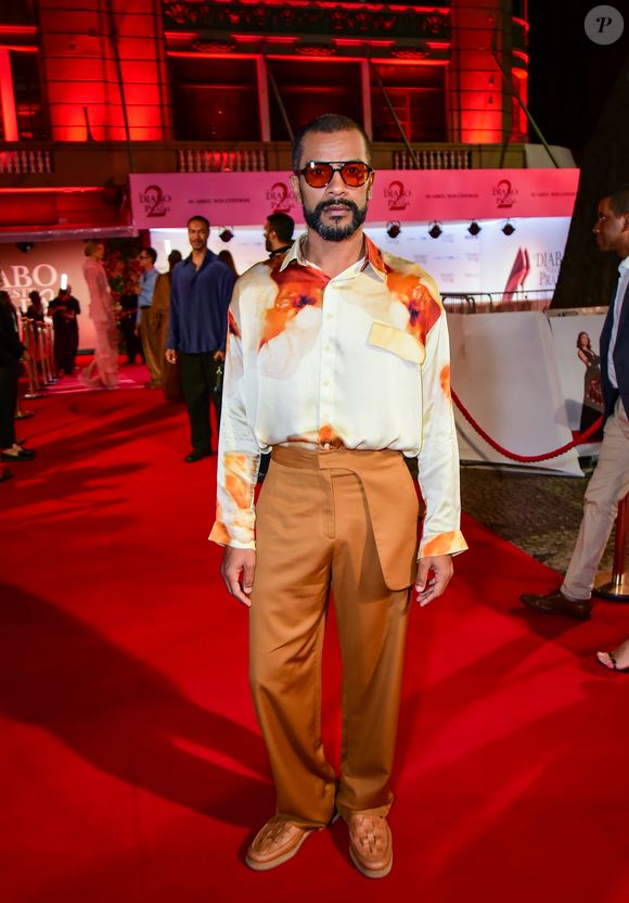 Samuel de Assis trouxe uma proposta de alfaiataria contemporânea e fluida. Ele vestiu uma camisa de seda com estampa abstrata/floral em tons terrosos e fundo off-white, com modelagem oversized. A calça de cintura alta, em tom caramelo acetinado, apresenta um detalhe de sobreposição no cós (estilo aba). Nos pés, ele fugiu do óbvio com sapatos de couro trançado (huaraches) e completou o visual com óculos de lentes translúcidas alaranjadas, mantendo a paleta quente