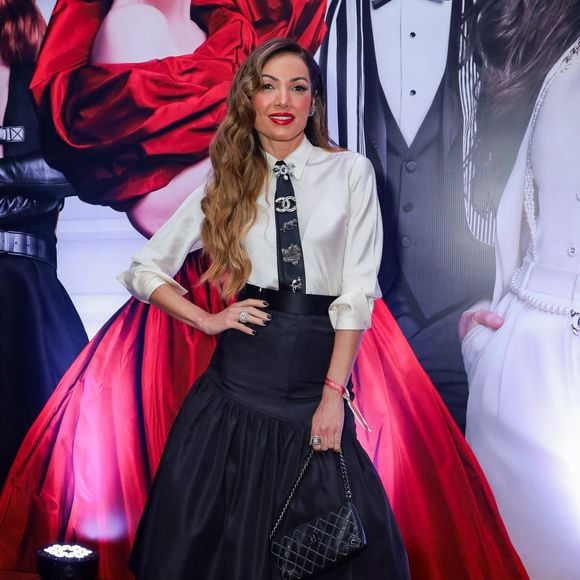 Patrícia Poeta apostou em uma estética 'preppy chic' com referências diretas à alta costura clássica. O conjunto é composto por uma camisa de seda off-white com mangas levemente bufantes e punhos ajustados, combinada a uma saia midi preta de tafetá com volume estruturado e cintura alta marcada por um cinto largo. O grande destaque da produção é a gravata preta estreita, adornada com diversos broches de luxo, incluindo o icônico logo da Chanel e elementos em pedraria