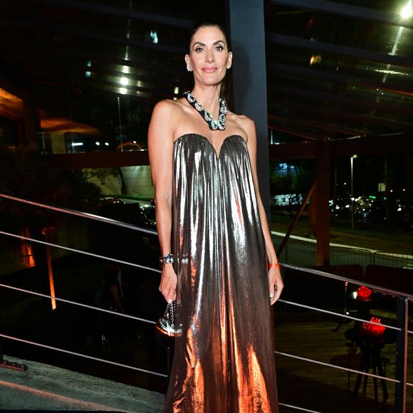 Isabela Fiorentino: veste um vestido longo sem alças (tomara que caia) em tecido metálico plissado em tom bronze/prateado, de modelagem evasê e caimento fluido. Como acessório de destaque, usa um colar imponente em formato de maxicorrente prateada e preta, além de pulseira metálica e uma clutch esférica