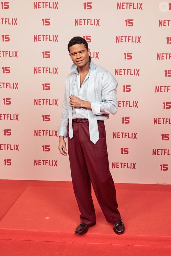 Gui Ferraz: veste uma camisa acetinada em tom azul-claro com gola alta desestruturada e faixas soltas no peito, combinada com calça de alfaiataria em tom vinho, cinto trançado e mocassim preto