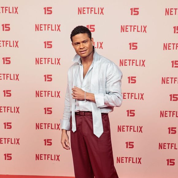 Gui Ferraz: veste uma camisa acetinada em tom azul-claro com gola alta desestruturada e faixas soltas no peito, combinada com calça de alfaiataria em tom vinho, cinto trançado e mocassim preto