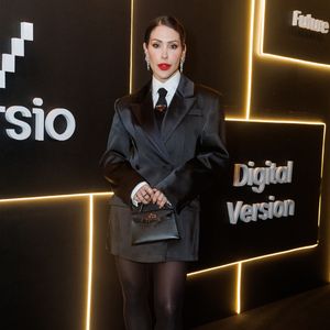 Anabela Falcone aposta em uma estética de alfaiataria desconstruída e de forte inspiração tomboy. Ela veste um blazer oversized de cetim preto estruturado, sobreposto a uma camisa social branca perfeitamente engomada e uma gravata preta fina, adornada com o clássico broche triangular da grife Prada no nó. Na parte inferior, o visual ganha ares de minivestido, complementado por meia-calça preta translúcida e sapatos de salto alto com bico fino envernizados
