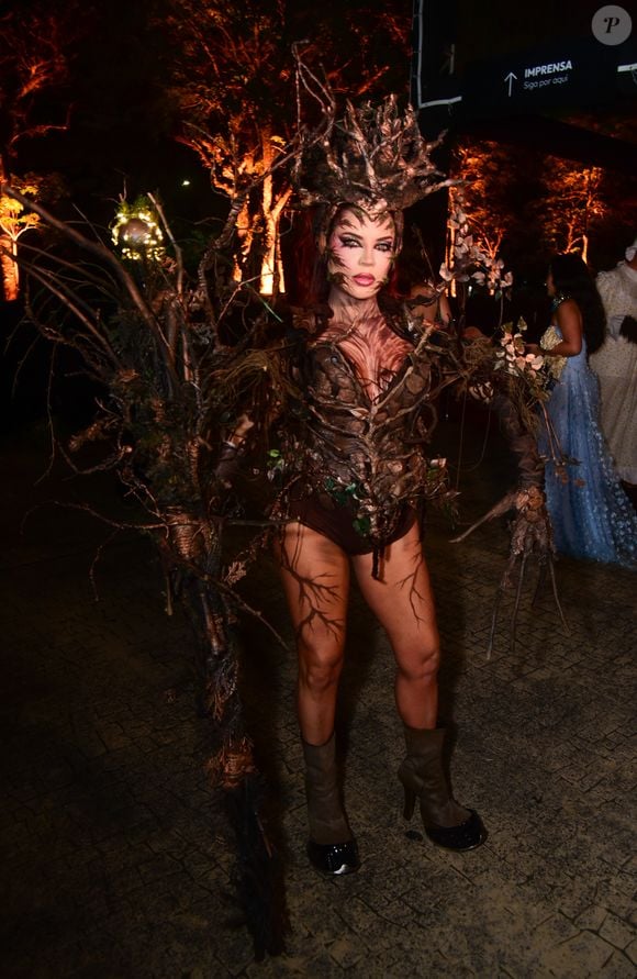 Gkay aposta em um traje conceitual inspirado na natureza, mimetizando uma árvore. O visual é composto por um corpete e top estruturado que simula textura de casca de árvore e raízes entrelaçadas em tons de marrom. Ela usa um adereço de cabeça volumoso e orgânico feito de galhos secos e ramagens, que se estende para uma estrutura de costas dramática. A pele visível, incluindo pernas e braços, está coberta por pintura corporal imitando veios de raízes e textura de madeira, finalizado com botas de cano curto marrons e maquiagem artística facial