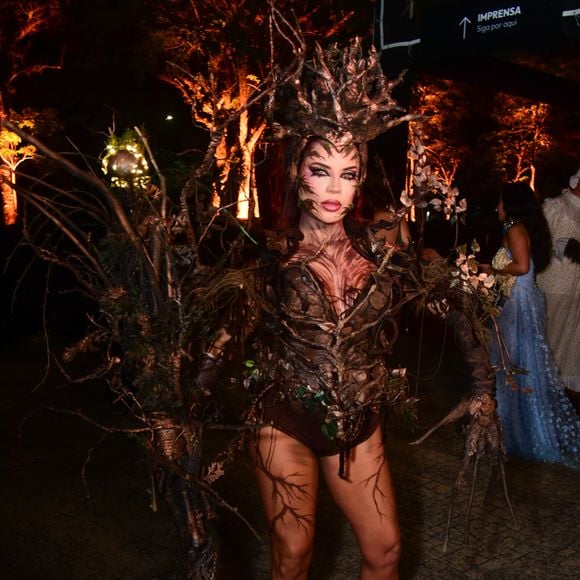 Gkay aposta em um traje conceitual inspirado na natureza, mimetizando uma árvore. O visual é composto por um corpete e top estruturado que simula textura de casca de árvore e raízes entrelaçadas em tons de marrom. Ela usa um adereço de cabeça volumoso e orgânico feito de galhos secos e ramagens, que se estende para uma estrutura de costas dramática. A pele visível, incluindo pernas e braços, está coberta por pintura corporal imitando veios de raízes e textura de madeira, finalizado com botas de cano curto marrons e maquiagem artística facial
