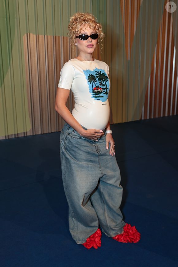 A criadora de conteúdo Malu Borges, conhecida por seus looks experimentais, exibiu sua gravidez com um visual que une o pop ao surrealismo. Ela utilizou uma t-shirt de látex branco da marca Normando, com uma estampa tropical centralizada, ajustada ao corpo para destacar a silhueta da gestação. A parte inferior foi composta por uma saia jeans de modelagem extremamente ampla e desestruturada, criando um jogo de proporções exageradas. Nos pés, as famosas sapatilhas de flores vermelhas tridimensionais adicionaram a textura e o volume que são marcas registradas de seu estilo autêntico