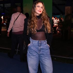Aline Fanju combinou casualidade e sofisticação em um look moderno. Ela vestiu uma blusa preta de tule transparente com gola alta, revelando um sutiã de renda por baixo, sobreposta por um blazer preto clássico. A produção se completou com calça jeans baggy de lavagem clara e sapatos mocassim tratorados em couro preto com fivela dourada