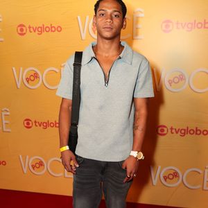 Apresenta um visual streetwear com camisa polo azul-claro texturizada e fecho em zíper, acompanhada de jeans cinza-escuro com lavagem estonada. Acessórios como bolsa tiracolo, relógio de pulseira branca e tênis pretos fecham a composição