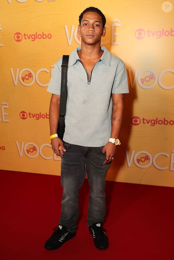 Apresenta um visual streetwear com camisa polo azul-claro texturizada e fecho em zíper, acompanhada de jeans cinza-escuro com lavagem estonada. Acessórios como bolsa tiracolo, relógio de pulseira branca e tênis pretos fecham a composição
