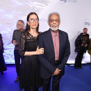 Gilberto Gil e Flora Gil: O cantor veste um terno azul-marinho clássico alinhado a uma camisa social em tom rosa-antigo de padronagem sutil, sem gravata. Flora acompanha com um vestido preto midi todo rendado em motivos florais, com transparência nos braços e colo, meia-calça e sapatos pretos