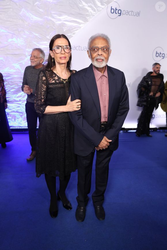 Gilberto Gil e Flora Gil: O cantor veste um terno azul-marinho clássico alinhado a uma camisa social em tom rosa-antigo de padronagem sutil, sem gravata. Flora acompanha com um vestido preto midi todo rendado em motivos florais, com transparência nos braços e colo, meia-calça e sapatos pretos