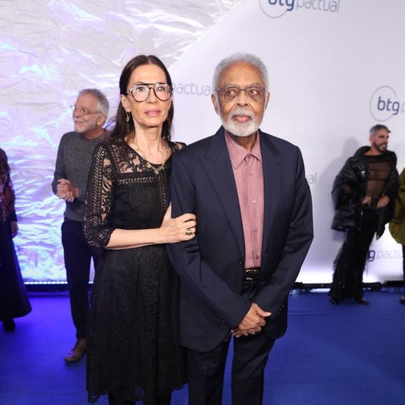 Gilberto Gil e Flora Gil: O cantor veste um terno azul-marinho clássico alinhado a uma camisa social em tom rosa-antigo de padronagem sutil, sem gravata. Flora acompanha com um vestido preto midi todo rendado em motivos florais, com transparência nos braços e colo, meia-calça e sapatos pretos