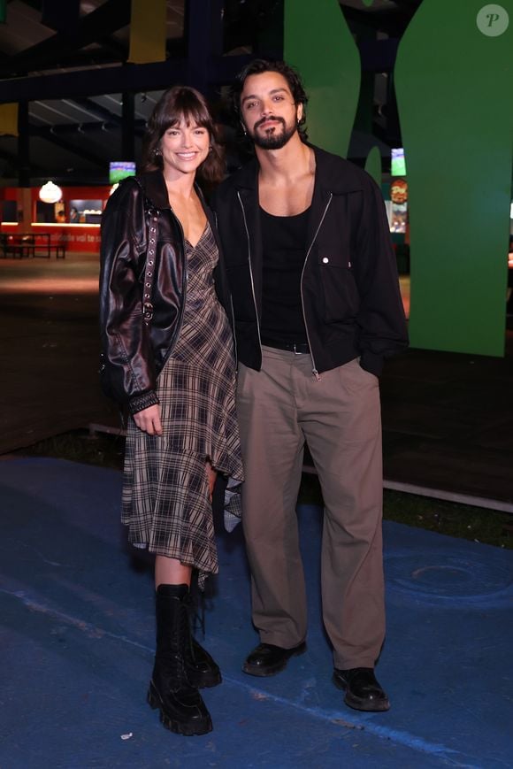 Agatha Moreira veste um vestido assimétrico com estampa xadrez em tons de marrom e bege, combinado com uma jaqueta de couro preta oversized e robustas botas tratoradas de amarrar. Rodrigo Simas complementa o par com calça de sarja bege de modelagem solta, regata preta sob uma jaqueta leve em estilo bomber preta com zíper e sapatos sociais pretos tratorados