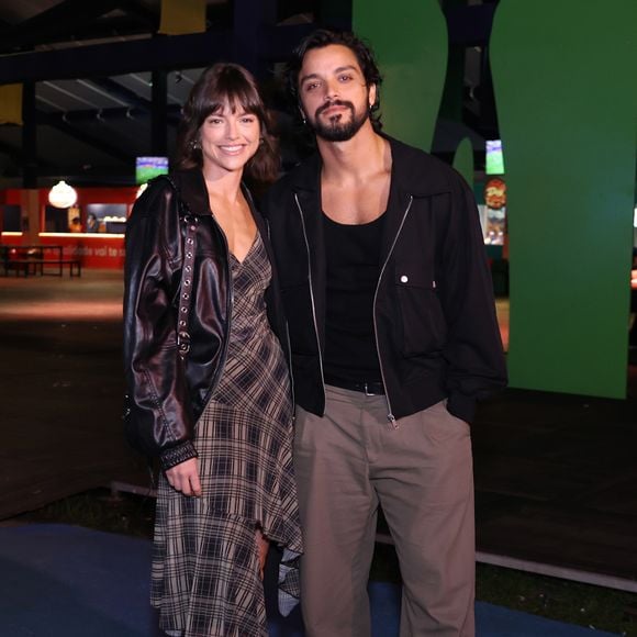 Agatha Moreira veste um vestido assimétrico com estampa xadrez em tons de marrom e bege, combinado com uma jaqueta de couro preta oversized e robustas botas tratoradas de amarrar. Rodrigo Simas complementa o par com calça de sarja bege de modelagem solta, regata preta sob uma jaqueta leve em estilo bomber preta com zíper e sapatos sociais pretos tratorados