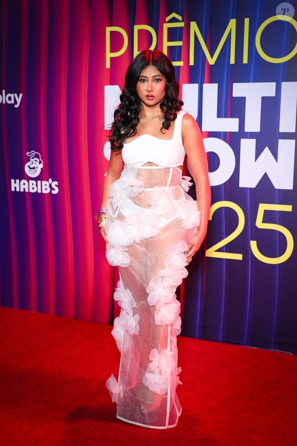 Zaynara apostou em um visual romântico com um toque moderno no Prêmio Multishow: ela surgiu com um conjunto todo branco formado por um top estruturado e uma saia longa transparente, adornada por grandes flores aplicadas em 3D que criam volume e movimento ao redor do corpo. O efeito é etéreo, quase de noiva fashionista, mas com um frescor jovial. O cabelo solto com ondas largas e a make iluminada completam o look, mantendo o clima leve e delicado da produção