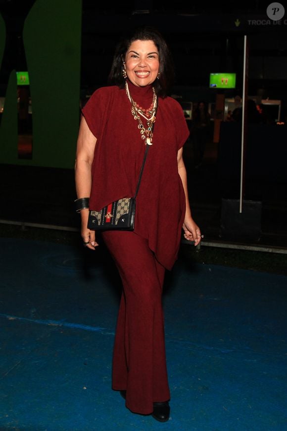 Fabiana Karla elege um conjunto monocromático texturizado em tom de vermelho-vinho (burgundy), formado por uma blusa de gola alta e caimento assimétrico sem mangas sobreposta a uma calça de corte flare no mesmo tecido. Como acessórios de destaque, usa um maximalista mix de colares de pedrarias e pérolas, além de uma bolsa transversal Gucci com estampa de monograma clássico