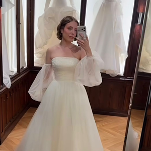 7. Princesa de tule com mangas bufantes soltas: vestido estilo princesa com corpete tomara que caia estruturado de acabamento liso e saia bem volumosa feita em camadas de tule. O visual é finalizado por mangas removíveis estilo ombro a ombro (off-the-shoulder), confeccionadas em organza/tule transparente com efeito bufante e punhos ajustados