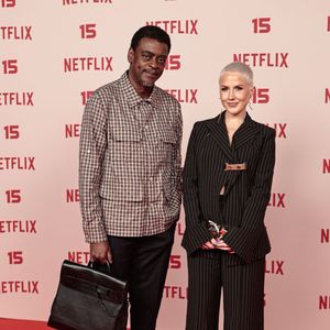 Seu Jorge: veste uma camisa de xadrez com bolsos frontais e calças escuras de corte reto, acompanhado por mocassins pretos e uma mala de mão estruturada em pele preta