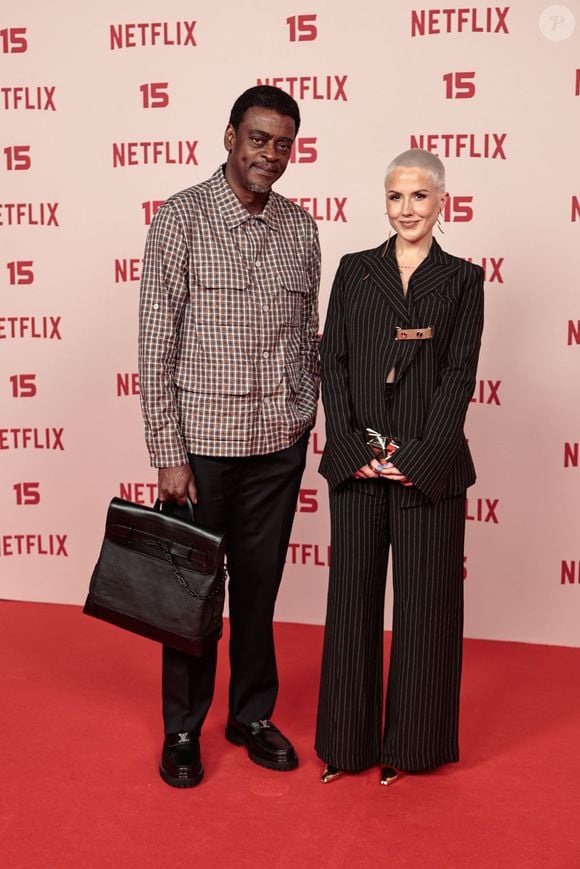 Seu Jorge: veste uma camisa de xadrez com bolsos frontais e calças escuras de corte reto, acompanhado por mocassins pretos e uma mala de mão estruturada em pele preta