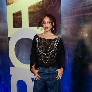 Livia Silva investiu em uma estética boho-chic com foco em texturas. Ela veste uma batinha preta de tecido leve e semitransparente, com mangas morcego e um decote canoa amplo. O peitoral da peça é ricamente decorado com um bordado maximalista em formato de escudo ou V, utilizando pedrarias em tons de bronze, branco e prata. A composição segue para uma linha urbana com calça jeans de modelagem oversized e lavagem escura, finalizada com sandálias de tiras finas e brincos longos com aplicações de conchas