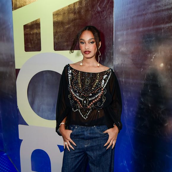 Livia Silva investiu em uma estética boho-chic com foco em texturas. Ela veste uma batinha preta de tecido leve e semitransparente, com mangas morcego e um decote canoa amplo. O peitoral da peça é ricamente decorado com um bordado maximalista em formato de escudo ou V, utilizando pedrarias em tons de bronze, branco e prata. A composição segue para uma linha urbana com calça jeans de modelagem oversized e lavagem escura, finalizada com sandálias de tiras finas e brincos longos com aplicações de conchas