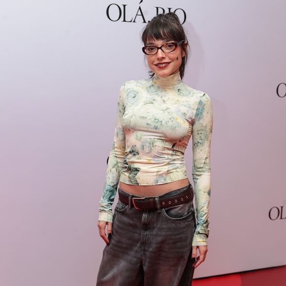 Bella Camero escolheu um visual que equilibra o romântico com o urbano. Na parte superior, utilizou uma blusa de gola alta e mangas longas com estampa floral delicada em tons de verde e creme, levemente transparente. O contraste ficou por conta da calça jeans de modelagem oversized em lavagem cinza estonada, presa por um cinto de couro marrom