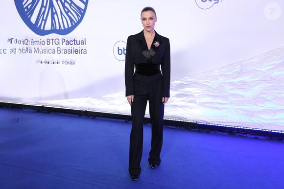 Antes de surgir com a criação inspirada na literatura brasileira, Alice também passou pelo tapete azul usando um elegante conjunto de alfaiataria preta. O visual era formado por blazer de lapela acetinada, cintura marcada por uma larga faixa de veludo, calça reta e um maxi broche colorido aplicado no peito, combinação que antecipava a sofisticação da noite