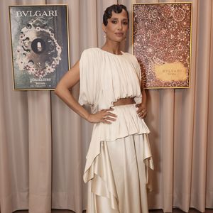 Camila Pitanga veste um conjunto de duas peças em seda na cor marfim ou off-white, composto por um top cropped intensamente plissado de corte assimétrico que deixa uma faixa do abdômen à mostra, e uma saia de cintura alta com sobreposições geométricas e pontas fluidas em cascata que criam movimento