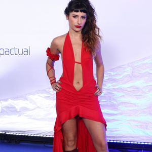 Duda Brack: vestido longo vermelho com decote frente única profundo que se estende em um recorte vazado até o umbigo e aberturas laterais na cintura. A saia possui drapeado frontal e modelagem high-low (curta na frente e longa atrás), combinada com botas de cano alto vinho repletas de fivelas metálicas