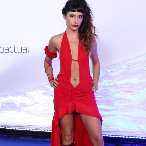 Duda Brack: vestido longo vermelho com decote frente única profundo que se estende em um recorte vazado até o umbigo e aberturas laterais na cintura. A saia possui drapeado frontal e modelagem high-low (curta na frente e longa atrás), combinada com botas de cano alto vinho repletas de fivelas metálicas